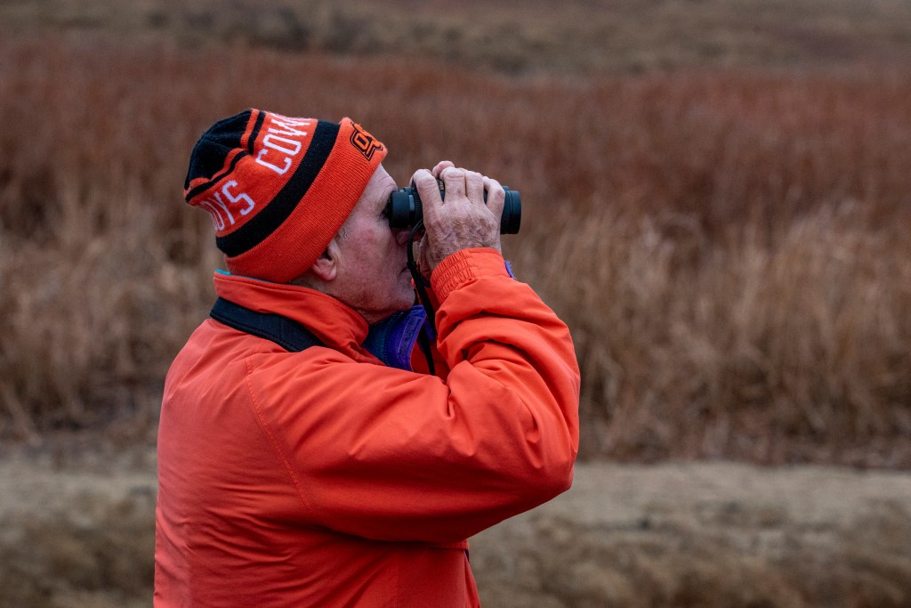 Profile of birder dressed in orange jacket and beanie looking through binoculars.